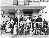 A Gathering of Norwegians at Spokkeli home in Camrose, Alberta, ca. 1912. L-R back row: Mrs. Edmund Thompson; Edmund Thompson; Lars Bjaaland family to right of verandah. L-R front row: I.Z. Hills; Mrs. I.Z. Hills; John Spokkeli; Myrtle Hills (child); Mrs. John Spokkeli; unknown; unknown; Mrs. John R. Lavick; Doctor John R. Lavick; next 5 unknown. Pastor Berg, extreme right, leaning on rail.