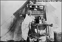 Round-up cook, southern Alberta, leaning on table attatched to back of wagon. Pies, meat, dishes shown. Note clock suspended. Date, 1905-1906.